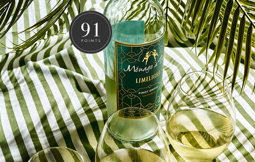Bottle and glasses of of Menage a Trois Limelight rated 91 points on a striped tablecloth with palm trees overhead