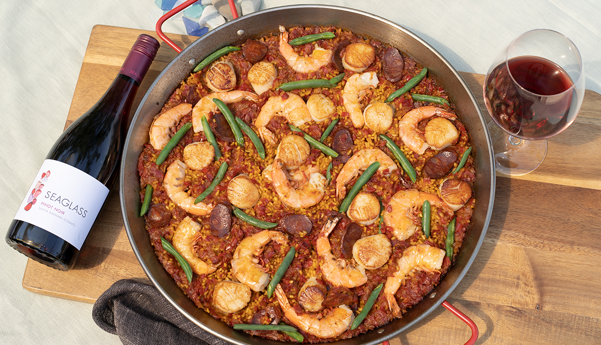 A large dish of Spanish Paella with loads of shrimp, scallops, and chrorizo, next to a glass and bottle of SEAGLASS pinot noir.