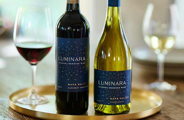 A bottle of Luminara alcohol-removed cabernet sauvignon and a bottle of Luminara alcohol-removed chardonnay on a gold charger set on a wooden table with glasses of filled wine nearby.