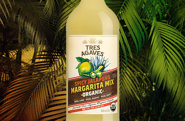 a bottle of Tres Agaves spicy jalapeno organic margarita mix with tropical plant leaves in the background.