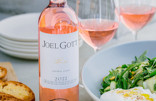 A bottle and two glasses of Joel Gott central coast rose next to crispy toasted bread, a stack of plates, and a bowl of salad.