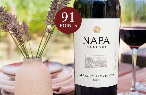 a bottle and glass of Napa Cellars cabernet sauvignon rated 91 points, next to a vase of purple flowers with green vineyards in the background.