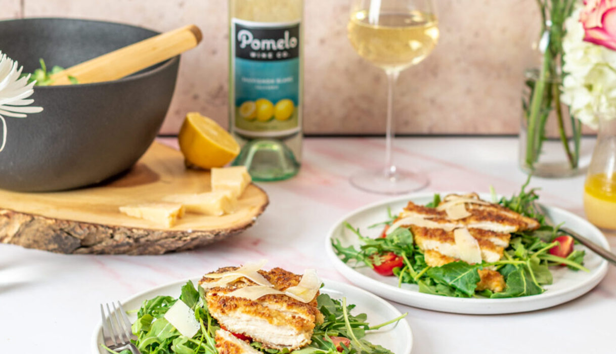 two plates of panko chicken salad with forks, with a bottle and glass of Pomelo sauvignon blanc and a salad bowl in the background with pink and white flowers nearby.